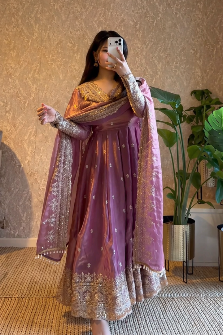Person wearing a traditional outfit with a purple and gold dress in a room with plants and a textured wall.
