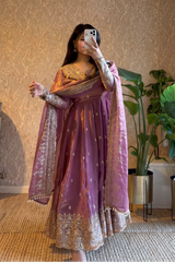 Person wearing a traditional outfit with a purple and gold dress in a room with plants and a textured wall.
