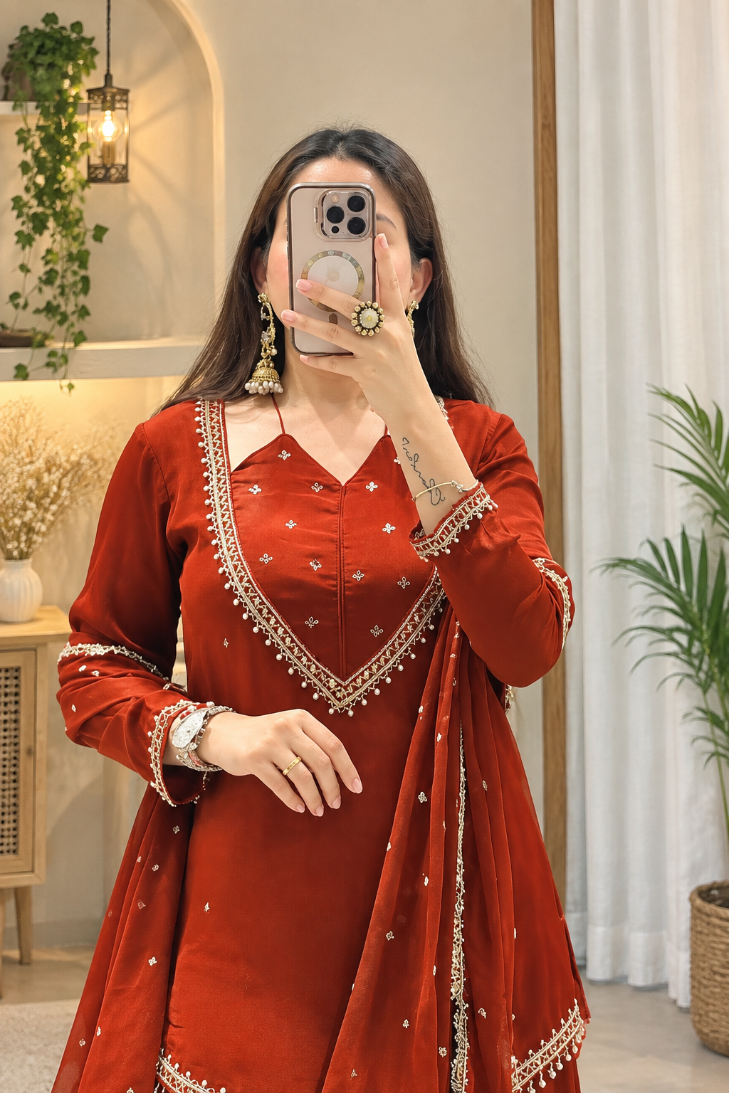 A woman takes a mirror selfie wearing a rust-red traditional Indian salwar kameez outfit with gold embroidery and a matching dupatta. The setting is a well-lit indoor space with plants and neutral walls.
