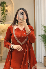 A woman takes a mirror selfie wearing a rust-red traditional Indian salwar kameez outfit with gold embroidery and a matching dupatta. The setting is a well-lit indoor space with plants and neutral walls.