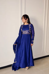 Woman wearing a blue traditional outfit with intricate designs against a white wall.