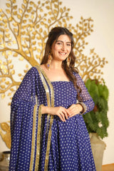 A woman wearing a navy blue traditional outfit with white printed motifs and gold border details, standing in front of a decorative gold tree wall piece and a potted plant.