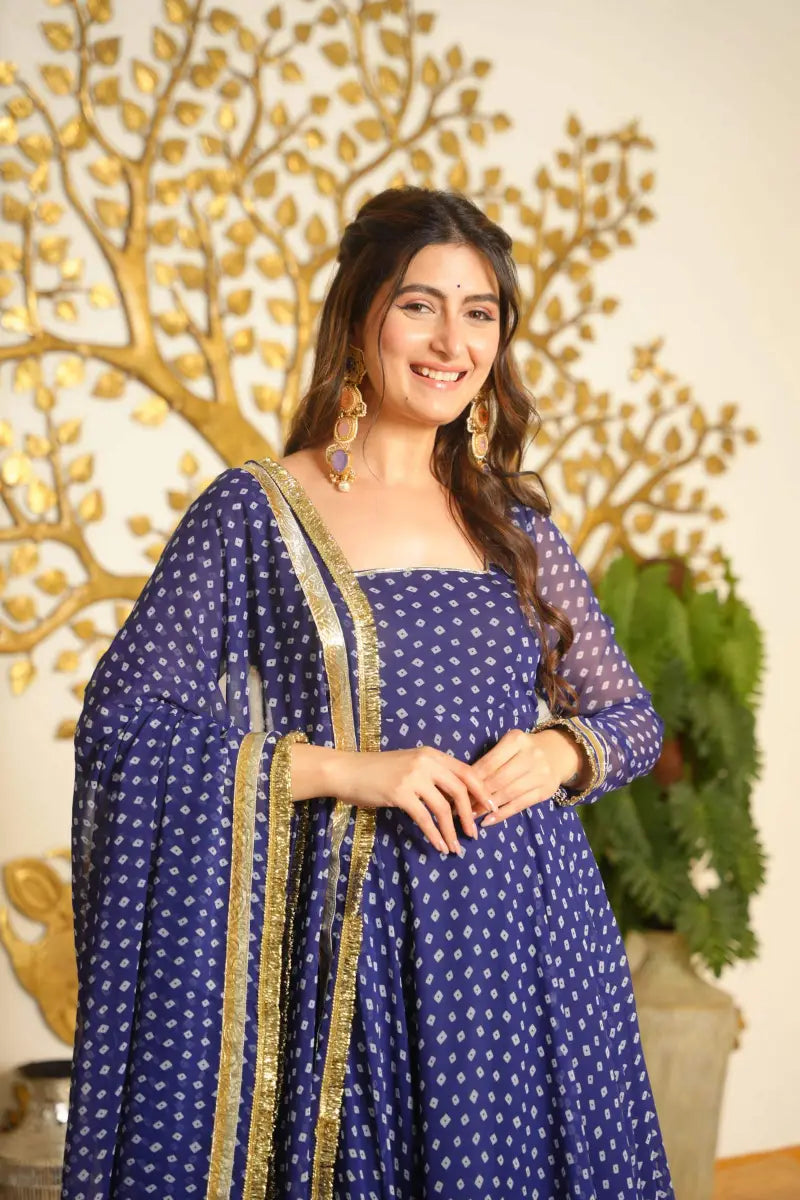 A woman wearing a navy blue traditional outfit with white printed motifs and gold border details, standing in front of a decorative gold tree wall piece and a potted plant.