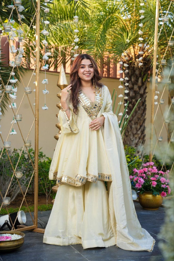 Woman in a cream traditional outfit standing outdoors with decorative plants and flowers.