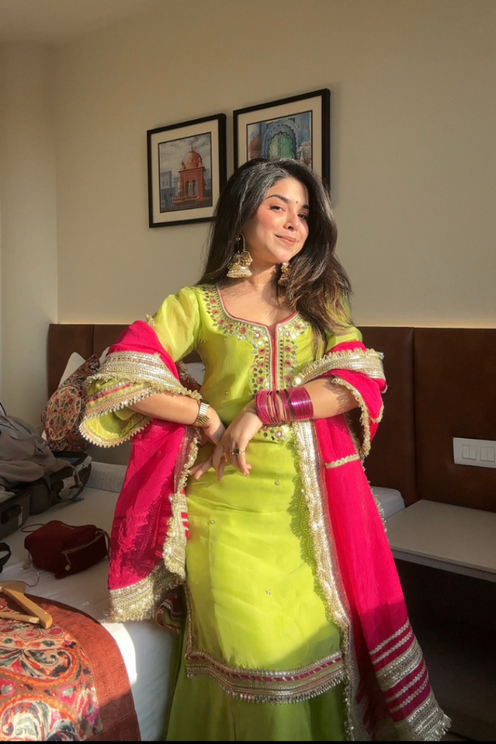 Woman in a traditional green and pink outfit standing in a room with framed pictures on the wall.