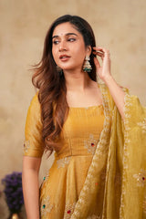A woman wearing a mustard yellow traditional outfit with matching embroidered dupatta and dangling earrings, posed against a warm beige textured backdrop.