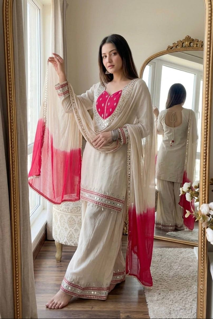A woman models a traditional Indian salwar kameez suit in cream with a vibrant pink heart-shaped chest embroidery and matching pink gradient dupatta, standing in front of a large gold-framed mirror in a room.