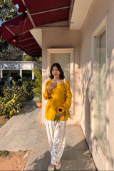 Woman in yellow top and white pants holding a sunflower, standing outside a building.
