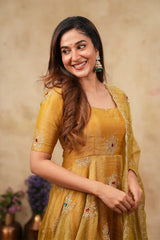 A woman smiling and wearing a mustard yellow embroidered traditional outfit with a matching sheer dupatta, set against a warm beige textured background.