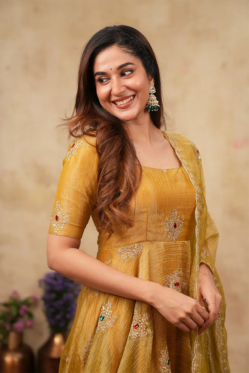 A woman smiling and wearing a mustard yellow embroidered traditional outfit with a matching sheer dupatta, set against a warm beige textured background.