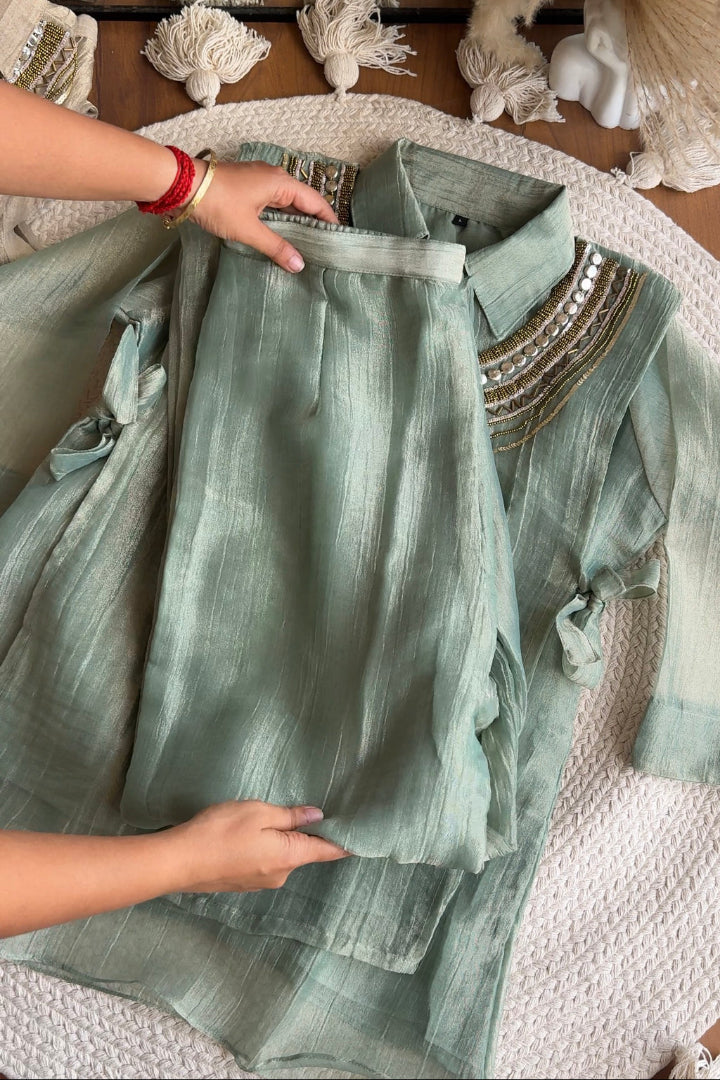 A sage green two-piece kurta set with a matching dupatta, laid out on a textured white blanket. A person's hands are visible holding the kurta to show its drape and texture.