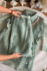 A sage green two-piece kurta set with a matching dupatta, laid out on a textured white blanket. A person's hands are visible holding the kurta to show its drape and texture.