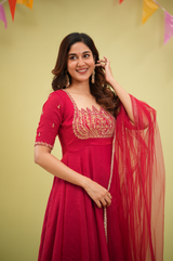A woman wearing a bright pink traditional Indian anarkali gown with gold embroidery and a matching sheer dupatta, posing against a pale yellow background with colorful hanging pennants.