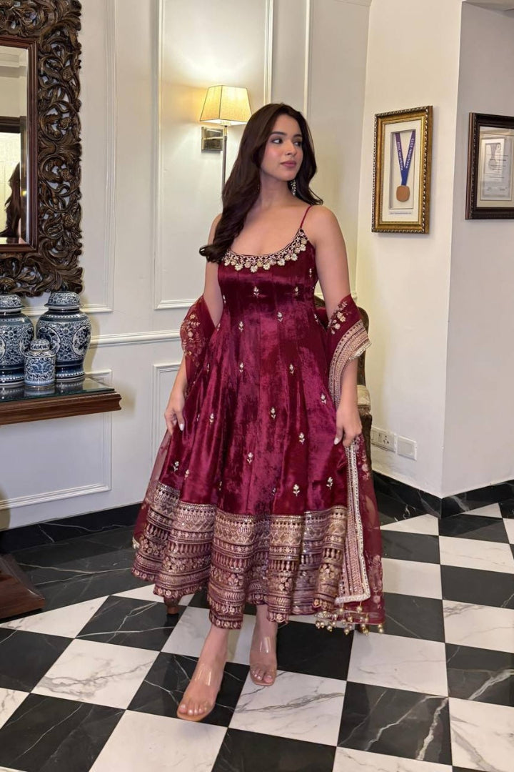 Woman in a maroon traditional outfit standing in a room with checkered floor and decorative elements.
