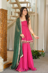 A woman models a bright pink three-piece salwar kameez set with white polka dots, featuring a matching green dupatta with silver borders, in an indoor setting with a wooden staircase.