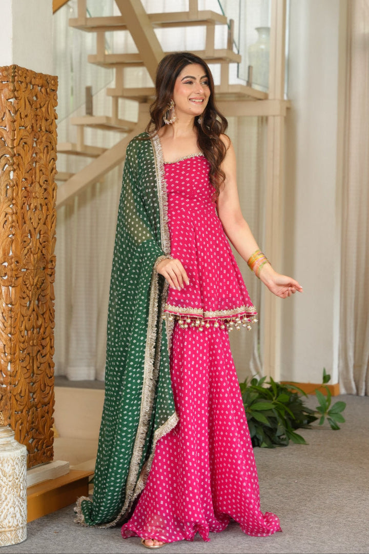 A woman models a bright pink three-piece salwar kameez set with white polka dots, featuring a matching green dupatta with silver borders, in an indoor setting with a wooden staircase.