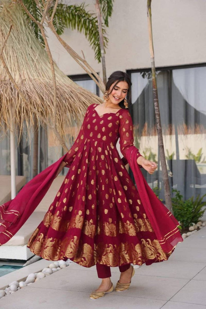 Woman in a red and gold traditional outfit standing outdoors with plants and a building in the background.