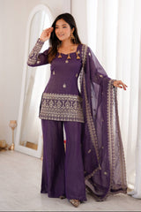 Woman in a purple traditional outfit with gold details standing in a bright room.