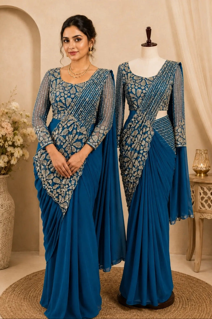 A woman and a mannequin display matching royal blue georgette sarees with intricate gold zari embroidery on the bodices. The setting features a warm beige background with a decorative arch, a vase of flowers, and a small side table.