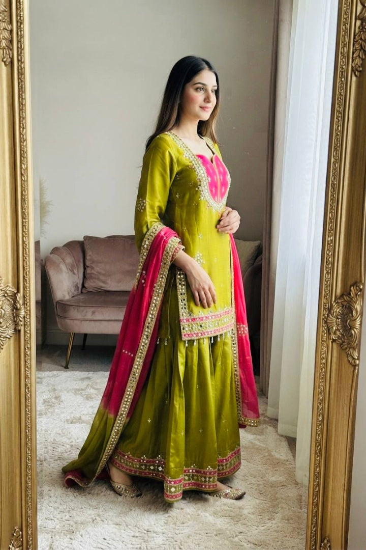 A woman stands in front of a large ornate gold mirror, wearing a traditional Indian outfit. The outfit consists of a bright lime green Anarkali kurta with pink embroidery on the neckline, a matching pink dupatta with gold borders, and a wide green sharara skirt with a pink and gold border.