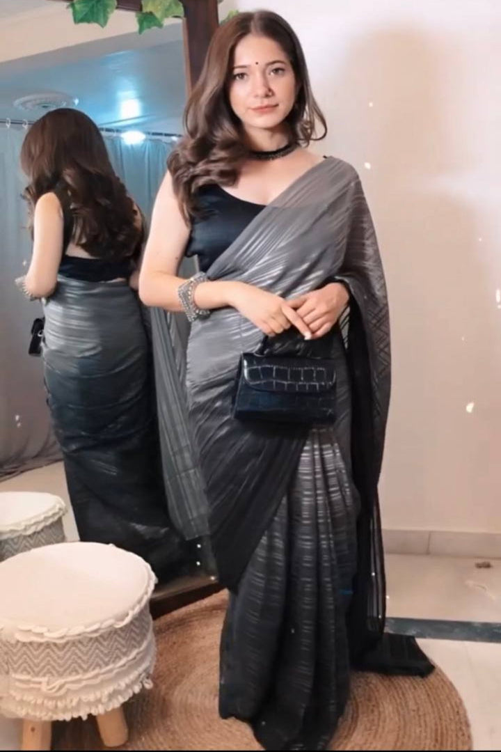 Woman in a black saree with a gray shawl holding a black handbag indoors.