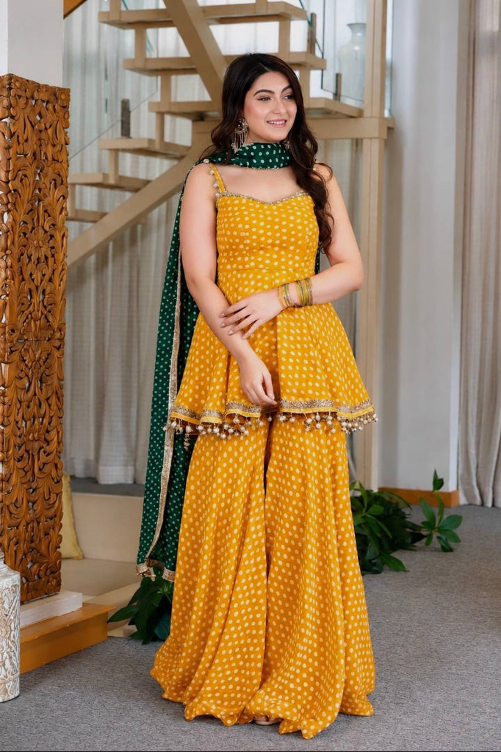 A woman models a yellow and white polka dot traditional Indian outfit with a green dupatta in an indoor setting with a staircase and curtains.