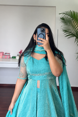 A woman takes a mirror selfie wearing a turquoise blue traditional dress with a matching dupatta and a light blue phone case. The dress features sheer embroidered sleeves, a keyhole cutout at the chest, and a central waist cutout.