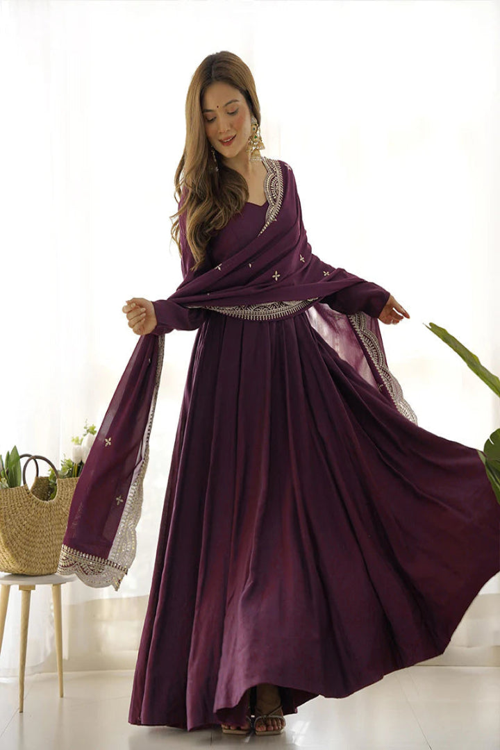 Woman wearing a long purple dress with white embroidery in a bright room.