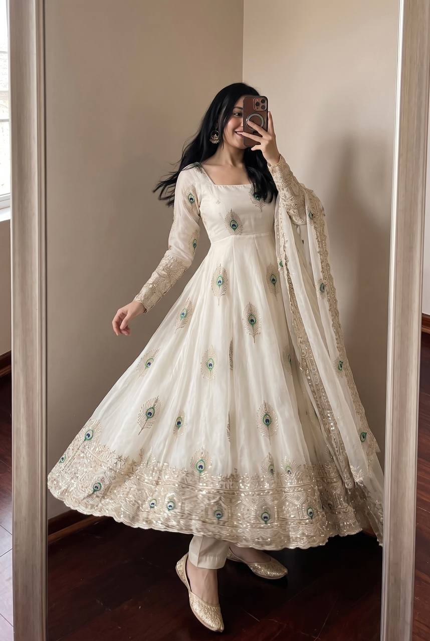 A woman takes a mirror selfie wearing a long-sleeved cream/off-white Anarkali gown with intricate gold embroidery and scattered teal green floral accents, paired with a matching sheer dupatta.
