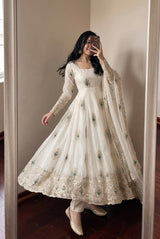 A woman takes a mirror selfie wearing a long-sleeved cream/off-white Anarkali gown with intricate gold embroidery and scattered teal green floral accents, paired with a matching sheer dupatta.