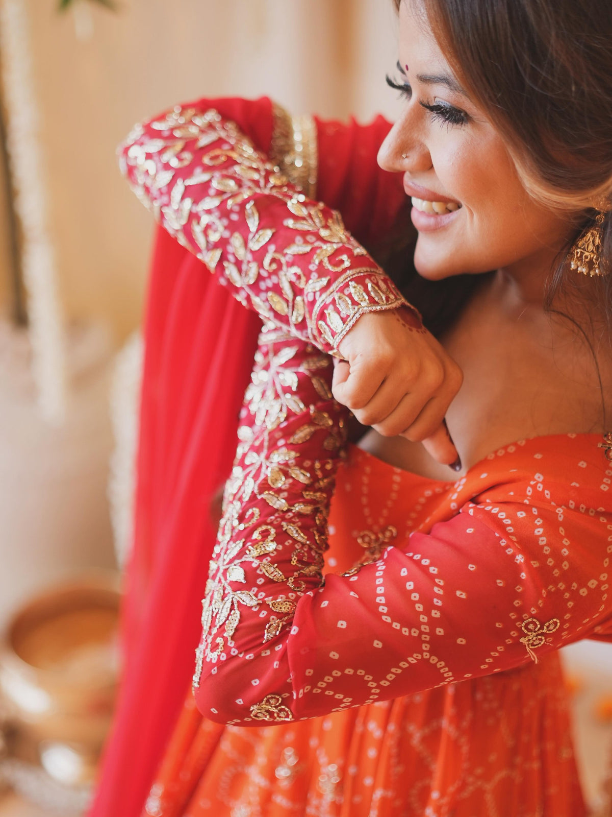 A woman wearing a vibrant red and orange traditional outfit with intricate gold embroidery and a matching red dupatta, captured in a warm, softly lit indoor setting.