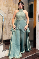 A woman wearing a sage green formal gown with a high-neck halter bodice, sheer embroidered overlay, and matching satin pants, standing in an indoor hallway with a staircase and elevator visible.