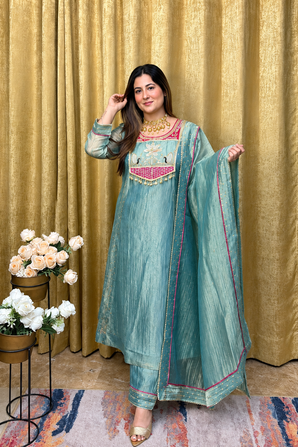 A woman models a traditional Indian outfit consisting of a teal blue-green kurta with pink and gold embroidery, matching trousers, and a sheer dupatta, set against a gold curtain backdrop with decorative flowers.