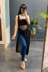 A woman takes a mirror selfie in a room wearing a black sleeveless tunic with colorful embroidered details at the waist, paired with blue skinny jeans.