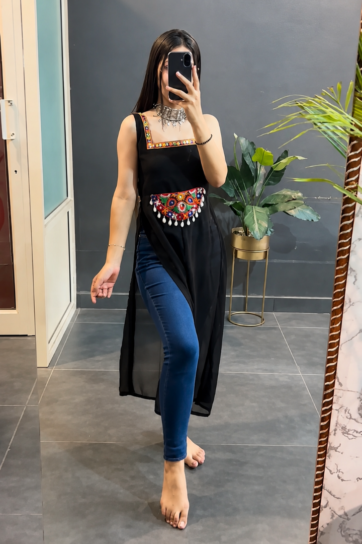 A woman takes a mirror selfie in a room wearing a black sleeveless tunic with colorful embroidered details at the waist, paired with blue skinny jeans.