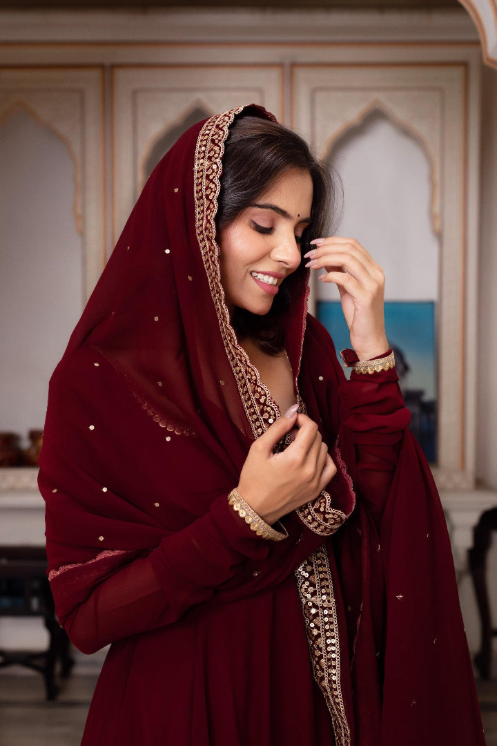 Woman wearing a maroon traditional outfit with gold embroidery indoors.