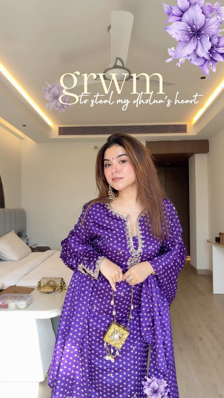 A woman wearing a purple and white polka dot kaftan dress with gold embroidery, standing in a modern bedroom setting. She is holding a small gold purse.
