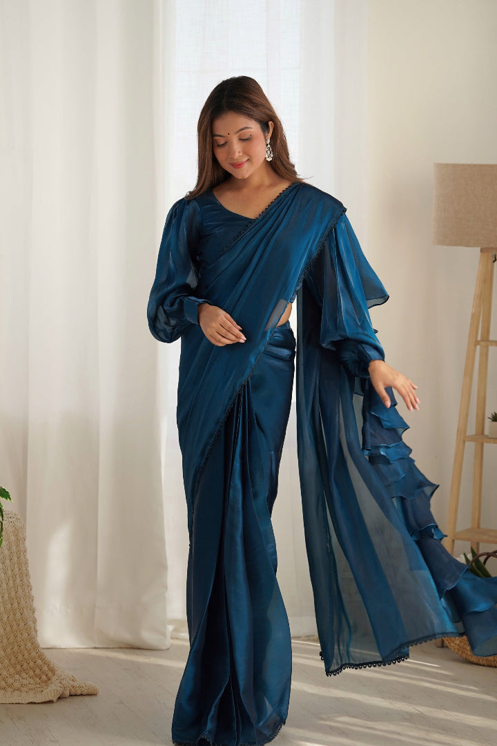 Woman wearing a blue saree standing in a room with white curtains and a lamp.