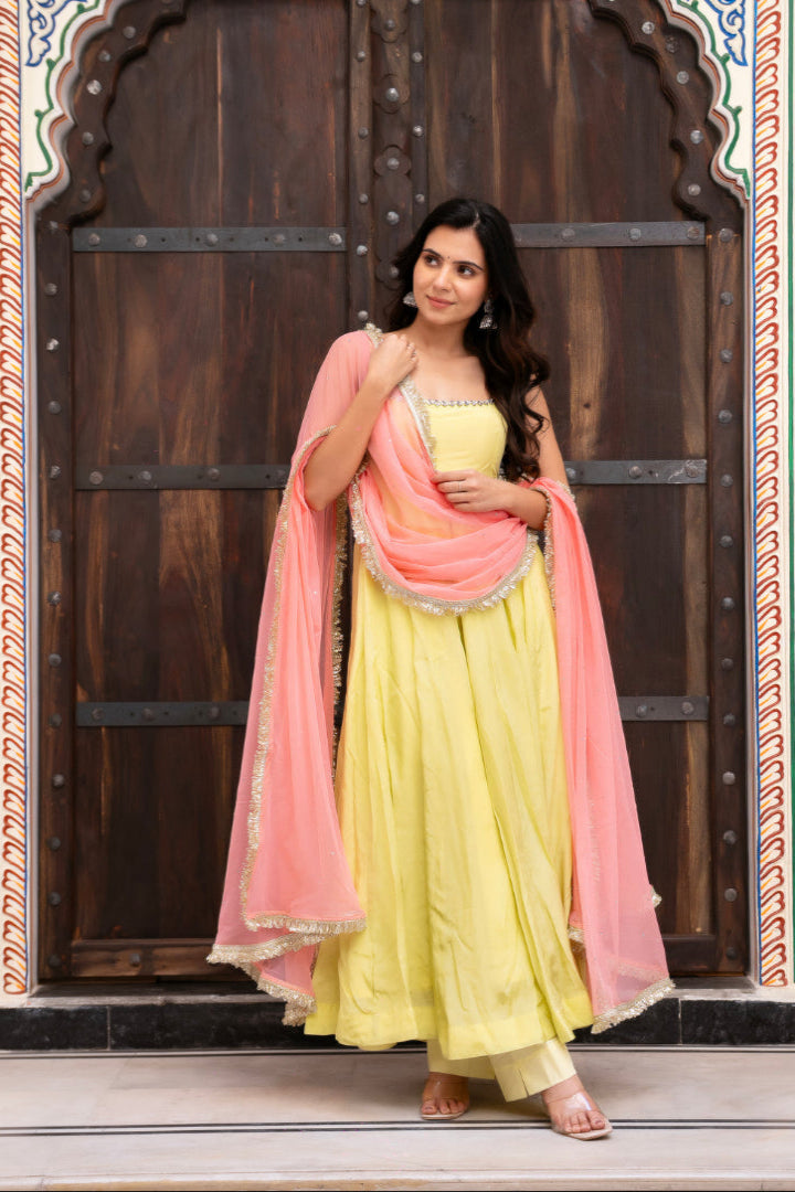 Woman in a yellow traditional outfit with a pink dupatta standing in front of an ornate wooden door.