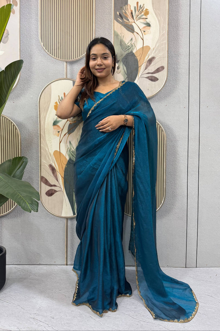 Woman in a blue saree standing indoors with decorative wall and plant in the background