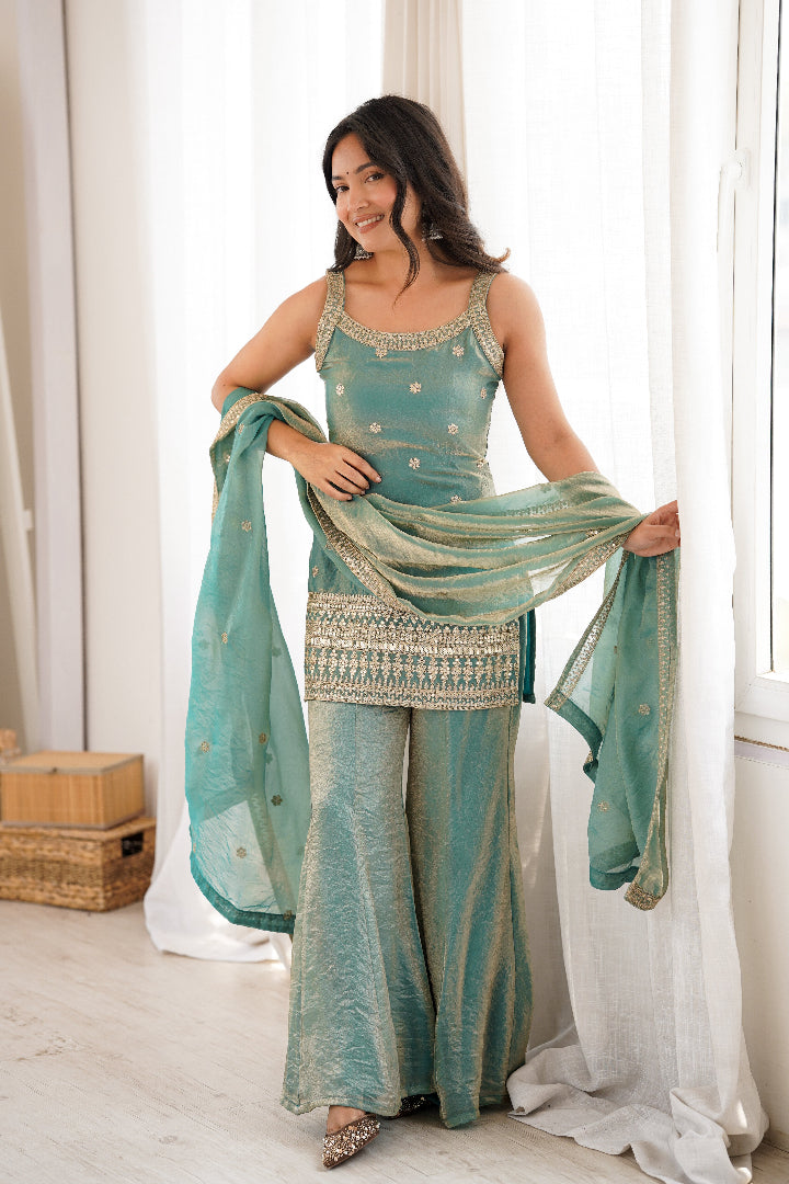 Woman in a teal traditional outfit with a matching dupatta, standing in a bright room.