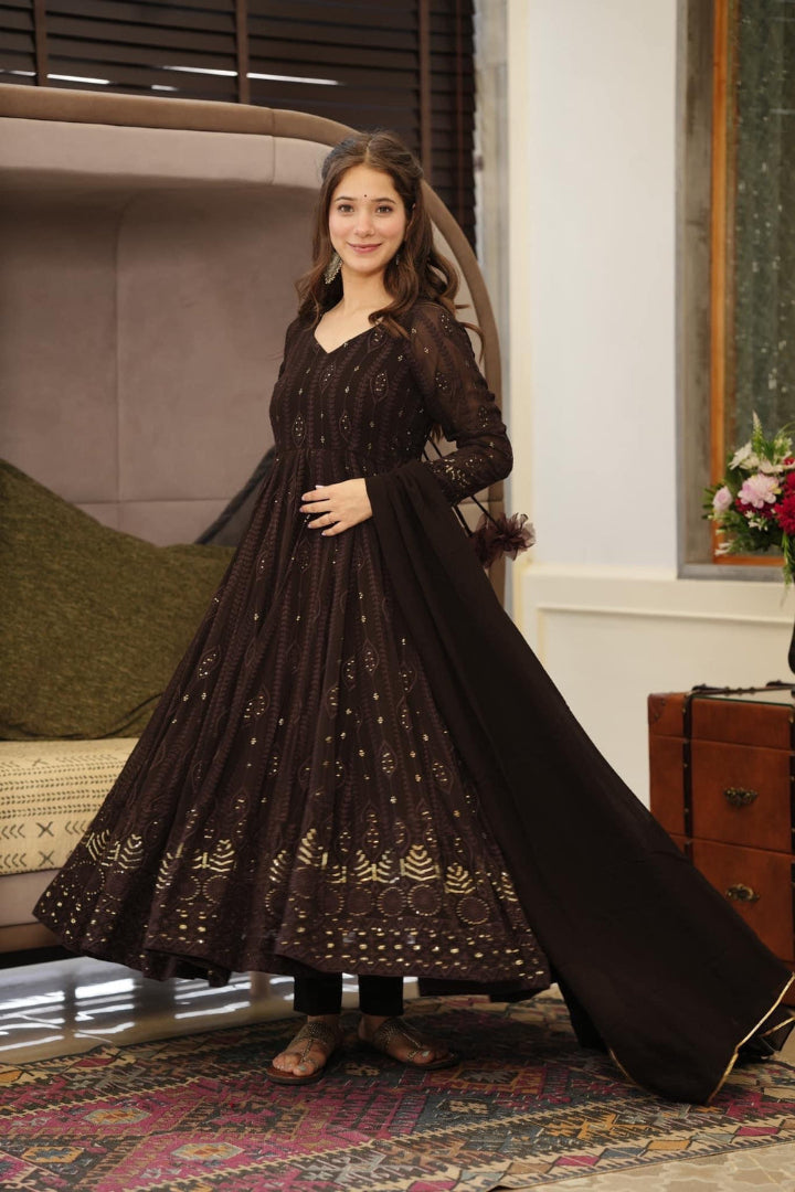 Woman in a long black dress with gold patterns standing in a room with a couch and decorative items.