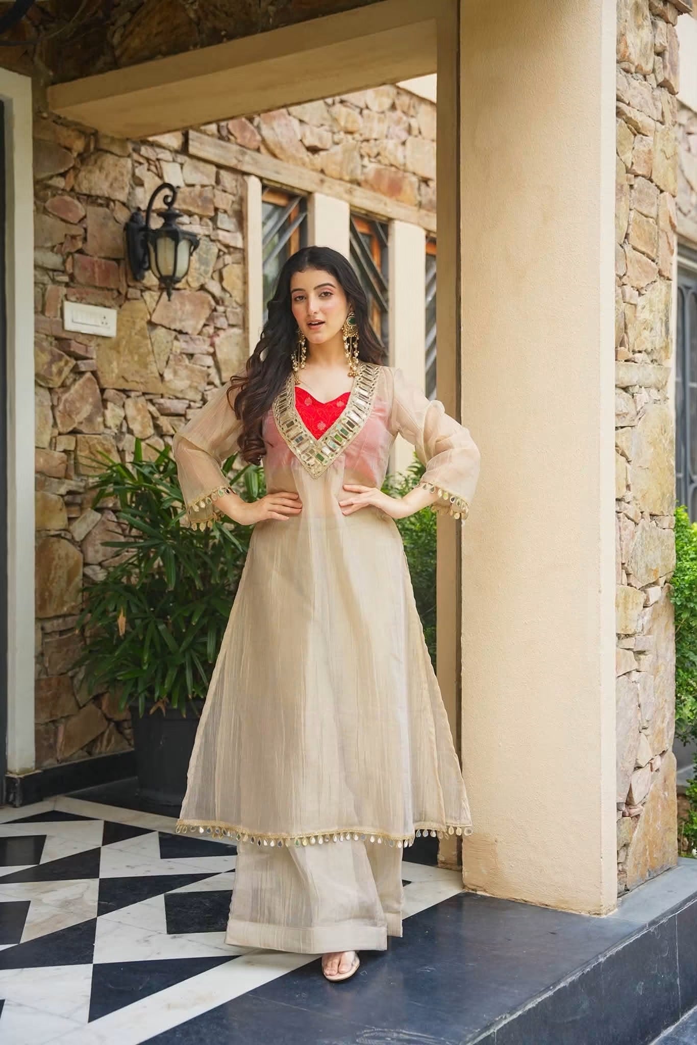 A woman models a beige Anarkali kurta with a red inner lining and gold embroidery, standing in a stone-walled doorway.