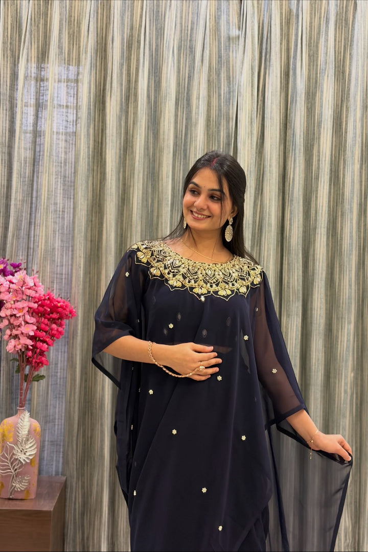 Woman in a dark blue traditional outfit with gold embroidery standing against a textured wall.