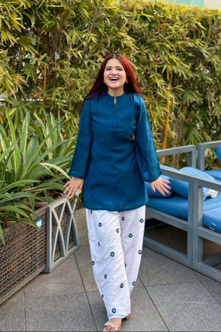 Woman in teal top and white pants with black patterns standing outdoors near plants and furniture.
