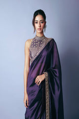 Woman wearing a purple saree with intricate designs against a plain background