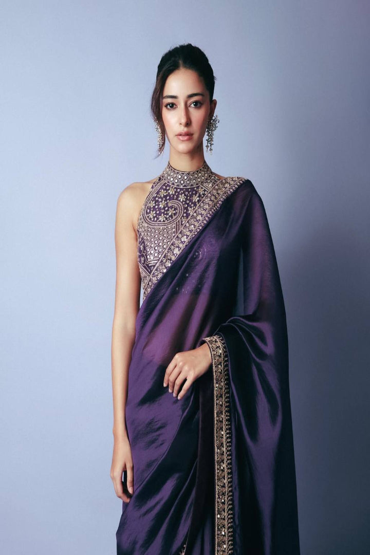 Woman wearing a purple saree with intricate designs against a plain background