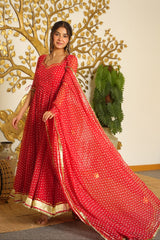 A woman models a red floor-length Anarkali gown with gold polka dots and a matching dupatta, set against a decorated indoor background with a gold tree mural.
