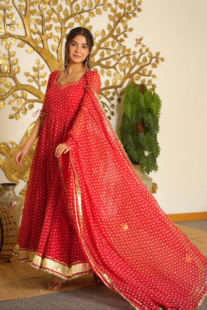 A woman models a red floor-length Anarkali gown with gold polka dots and a matching dupatta, set against a decorated indoor background with a gold tree mural.