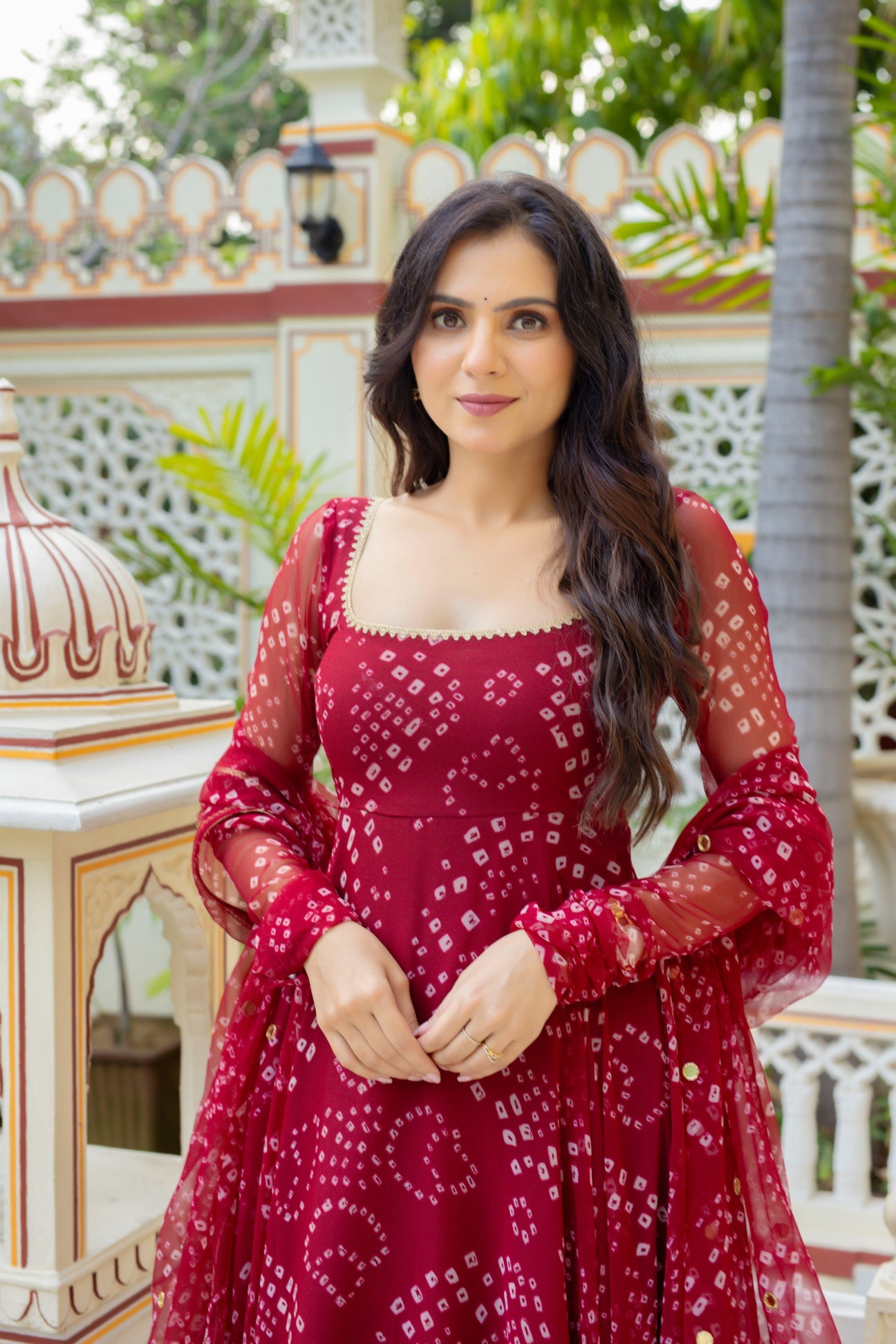 A woman wearing a deep red Anarkali gown with white bandhani tie-dye patterns, standing in front of a traditional Indian courtyard background.